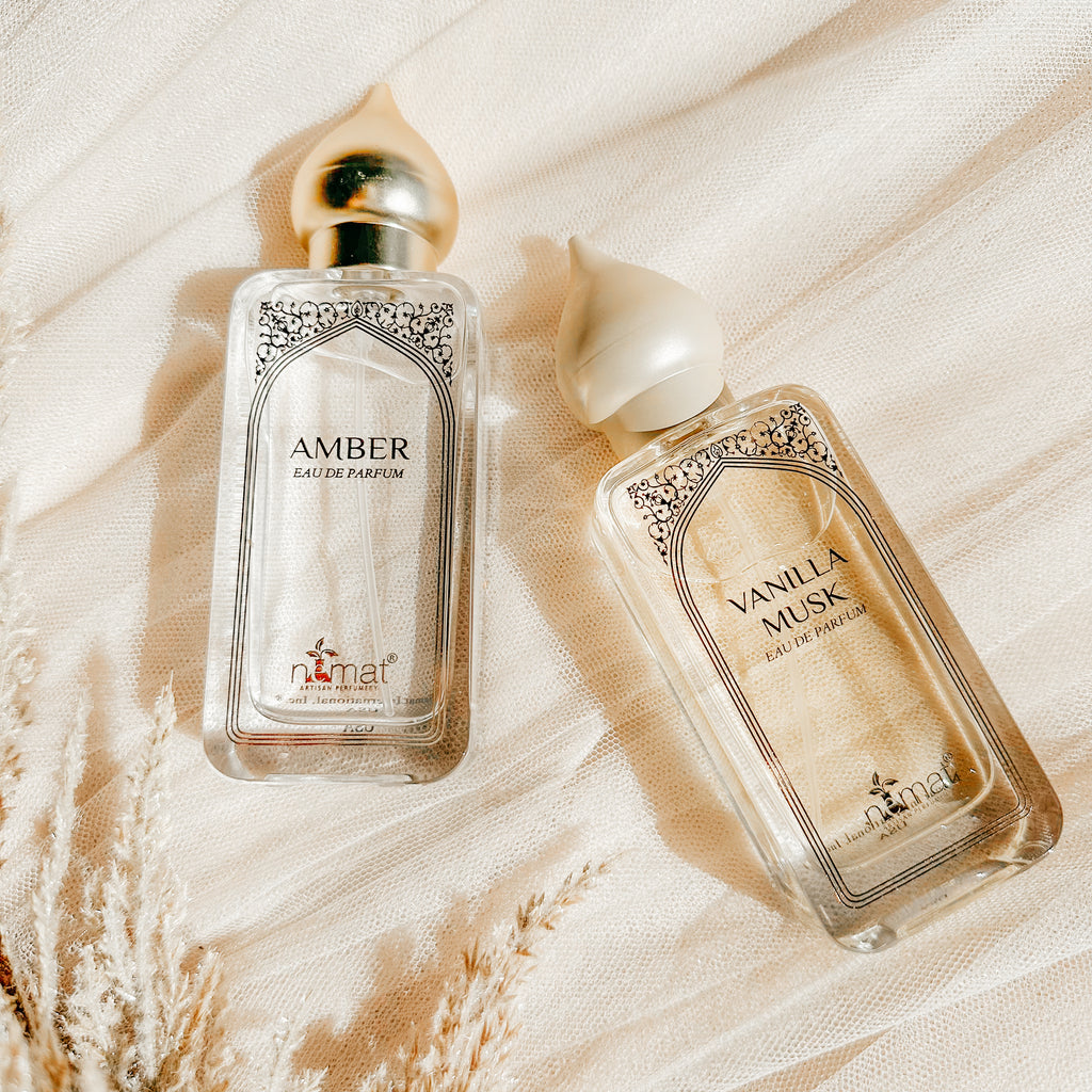 Two perfume bottles, one labeled 'AMBER' and the other 'VANILLA MUSK', are presented on a soft, textured background with a sheen, suggesting luxury and elegance.